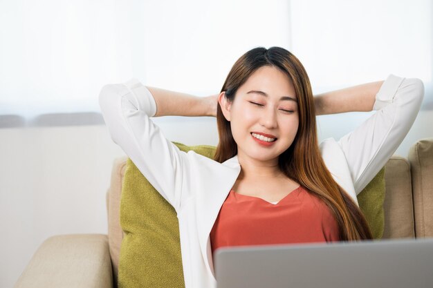 Mulher de negócios asiáticos jovem sentado relaxando no sofá na sala de estar em casa trabalhando com laptop.