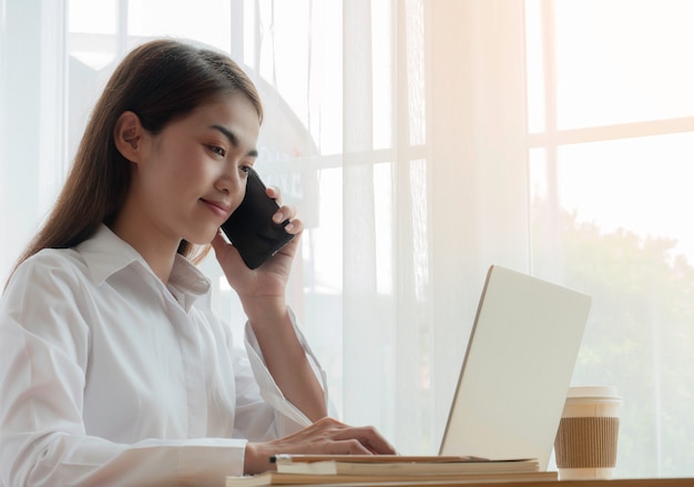 Mulher de negócios asiáticos jovens falando telefone inteligente e trabalhando com labtop