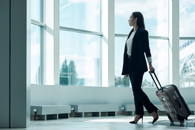 Mulher de negócios asiáticos jovens no aeroporto com saco de carrinho de bagagem, à espera de partida