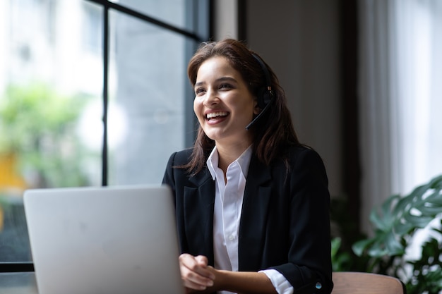 Mulher de negócios atraente asiática em ternos e fones de ouvido sorrindo enquanto trabalha com o computador