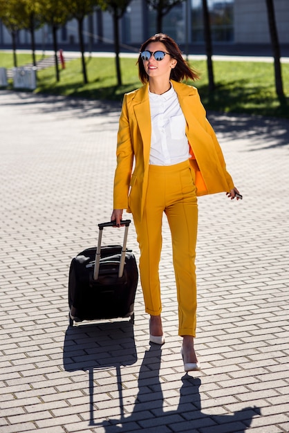 Mulher de negócios atraente elegante terno amarelo puxa uma mala, apressa-se para uma reunião de negócios. mulher de negócios atraente indo em uma viagem de negócios, puxando sua mala ao longo da calçada atrás dela.