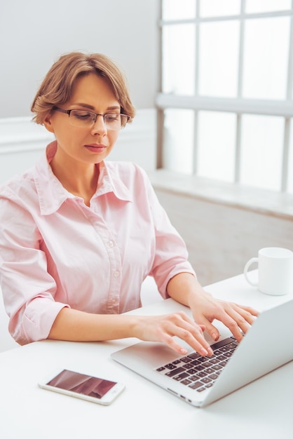 Mulher de negócios atraente em camisa clássica e óculos está usando um laptop enquanto trabalha no escritório