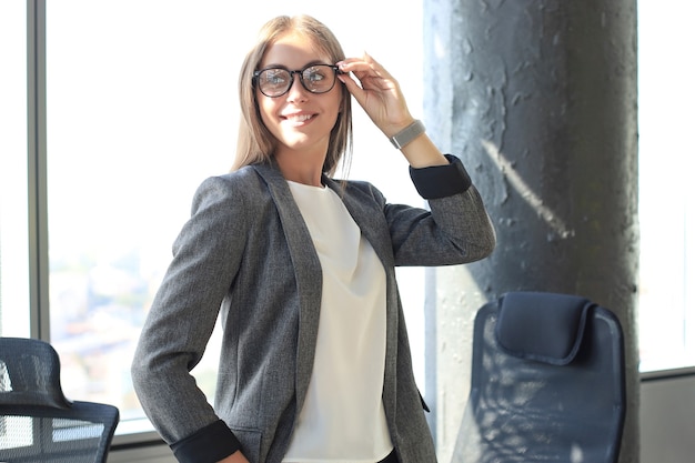 Mulher de negócios atraente sorrindo e segurando os óculos em pé no escritório.