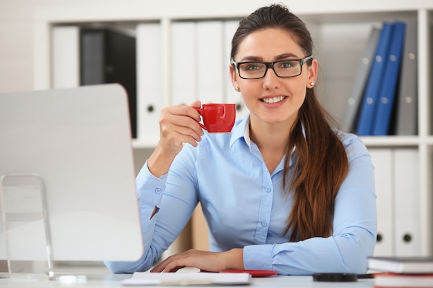 Mulher de negócios bebe café no escritório em uma mesa de uma caneca vermelha