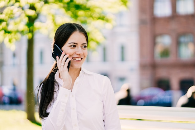 Mulher de negócios bem sucedido falando no celular enquanto caminhava ao ar livre, sorri amplamente aproveitando o bom dia. Mulher de negócios da cidade trabalhando.