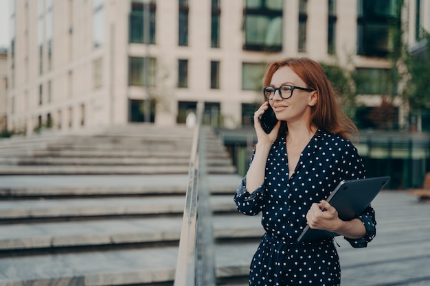 Mulher de negócios bonita confiante com laptop falando no celular enquanto caminha na rua da cidade,