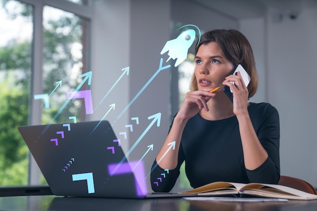 Mulher de negócios bonita e atraente pensativa com roupa formal trabalhando no laptop no local de trabalho do escritório em segundo plano Conceito de inicialização de uma nova tecnologia de negócios Holograma de ícone de foguete Falando telefone