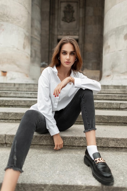 Mulher de negócios bonita e elegante em roupas de moda casual com camisa branca e jeans preto com sapatos senta-se nos degraus perto de um prédio antigo na cidade Moda e estilo de beleza