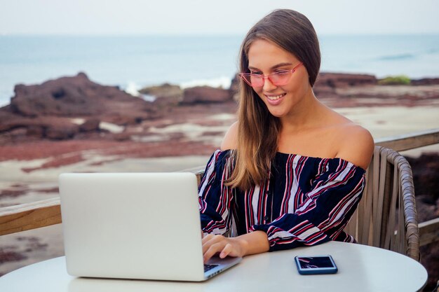 Mulher de negócios bonita e jovem em uma blusa listrada elegante com ombros abertos, sentado à mesa com um laptop no terraço do restaurante de verão na garota ocean.freelancer trabalhando online.