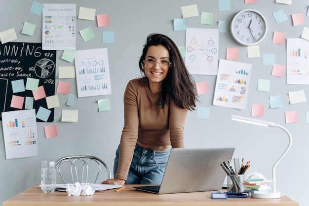 Foto mulher de negócios bonita e sorridente com óculos no escritório encostada na mesa