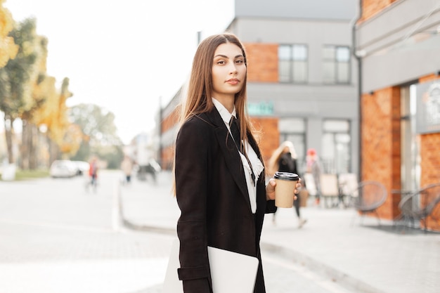 Mulher de negócios bonita elegante em roupas pretas extravagantes com um laptop e uma xícara de café andando na cidade Gerente profissional de rh feminino na rua