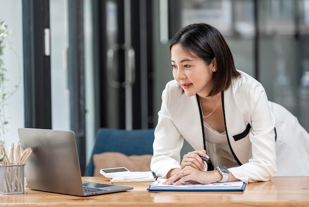 Mulher de negócios bonita feliz asiática em traje formal trabalhando no laptop no local de trabalho