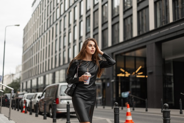 Mulher de negócios bonita na moda em roupas da moda preta com um vestido elegante e uma bolsa de couro com uma xícara de café caminha na cidade perto de um prédio de escritórios moderno Menina bonita urbana