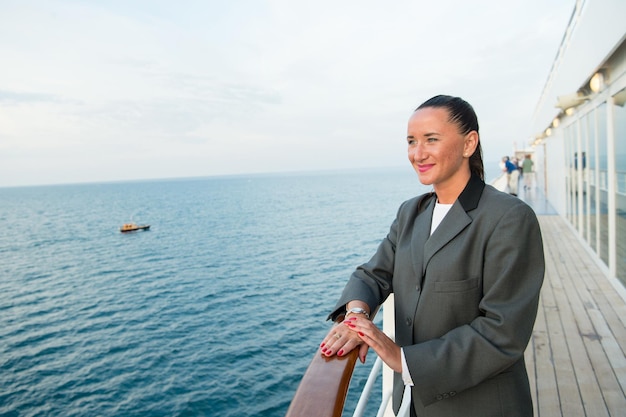 Mulher de negócios bonita ou garota bonita feliz com rosto sorridente em jaqueta cinza, manicure vermelha no convés do navio, olhando para o mar, a água do oceano com barco à vela no fundo do céu nublado, viagens e turismo