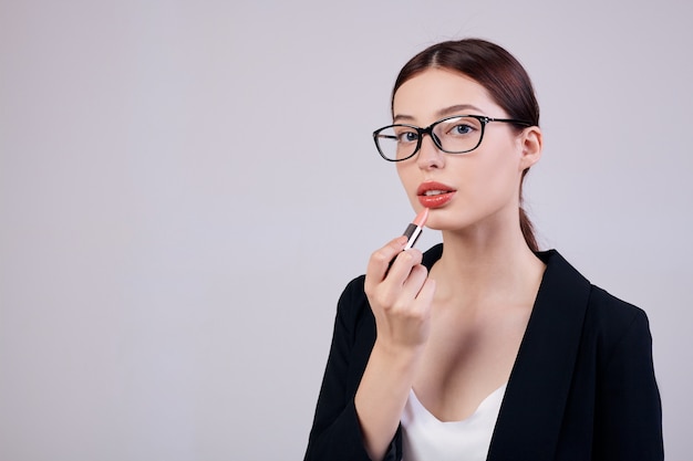 Mulher de negócios calmo de aparência agradável com batom rosa está de pé nas costas cinza em uma jaqueta preta, camiseta branca e óculos de computador. Trabalhando muito. Trabalhador ocupado.