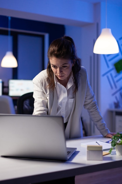 Mulher de negócios cansada e focada lendo e-mails no laptop em pé perto da mesa em uma empresa iniciante