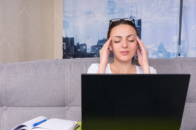 Mulher de negócios cansada fazendo uma massagem na cabeça enquanto trabalha em um laptop e faz seus negócios em casa, sentada em um sofá