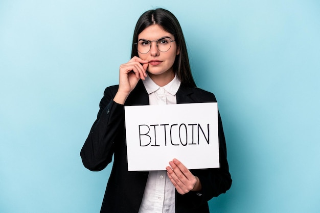 Mulher de negócios caucasiana jovem segurando um cartaz de bitcoin isolado em fundo azul com os dedos nos lábios, mantendo um segredo