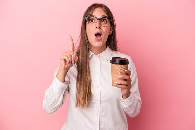 Mulher de negócios caucasiana jovem segurando um take away isolado no fundo rosa, apontando para cima com a boca aberta.