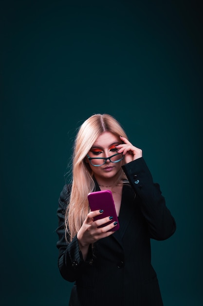 Mulher de negócios caucasiana jovem usando um telefone celular vestindo um blazer preto e óculos em um estúdio fotográfico com um fundo azul escuro.