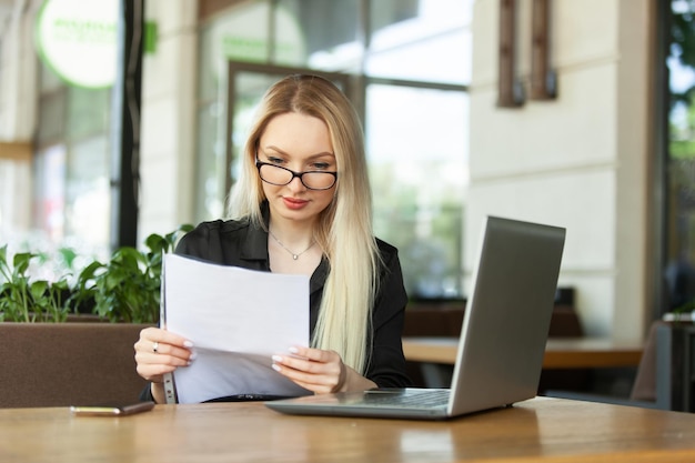 Mulher de negócios caucasiana loira examina folhas de documentos e se senta em uma mesa com um laptop em um café ao ar livre mulher de negócios moderna estilo de vida