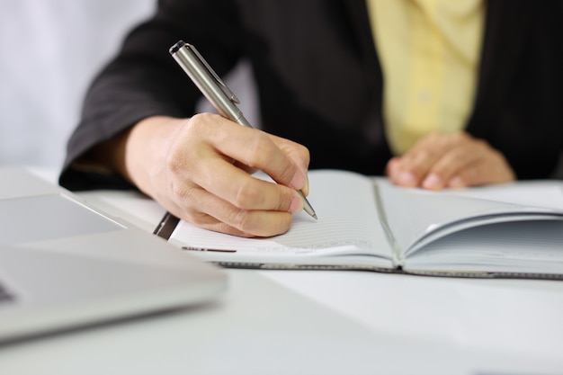 Mulher de negócios com as mãos em um terno preto sentado e escrevendo o caderno e trabalhando ou usando o computador na mesa branca com papelada