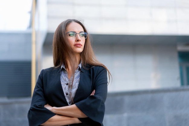 Mulher de negócios com cabelo comprido vestida de jaqueta preta óculos em pé ao ar livre perto do prédio de escritórios corporativos com as mãos postas Retrato de mulher de negócios caucasiano bonita na rua da cidade Vista lateral