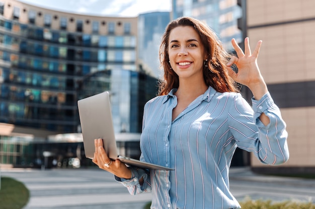 Mulher de negócios com camisa segura laptop e mostra sinal de ok ao ar livre