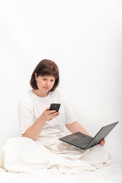Mulher de negócios com laptop e telefone na cama contra uma parede branca. Jovem trabalha sentada na cama. Moldura vertical
