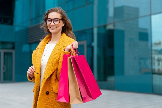 Mulher de negócios com sacolas de compras vestida de casaco amarelo caminhando ao ar livre superfície do edifício corporativo mulher de negócios caucasiano perto de prédio de escritórios venda de temporada elegante empresária