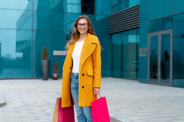 Mulher de negócios com sacos de compras vestido de casaco amarelo andando ao ar livre fundo de edifício corporativo Mulher de negócios caucasiana perto de prédio de escritórios Venda de temporada de empresária elegante