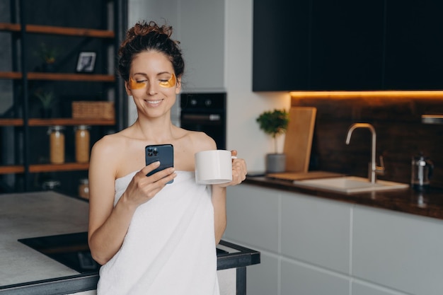 Mulher de negócios com skincare sob tapa-olhos lê notícias de mensagens no smartphone após o banho matinal