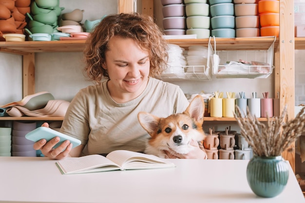 Mulher de negócios concentrada sorridente segurando smartphone com corgi de cachorro fofo na oficina Loja de pratos de bebê de silicone