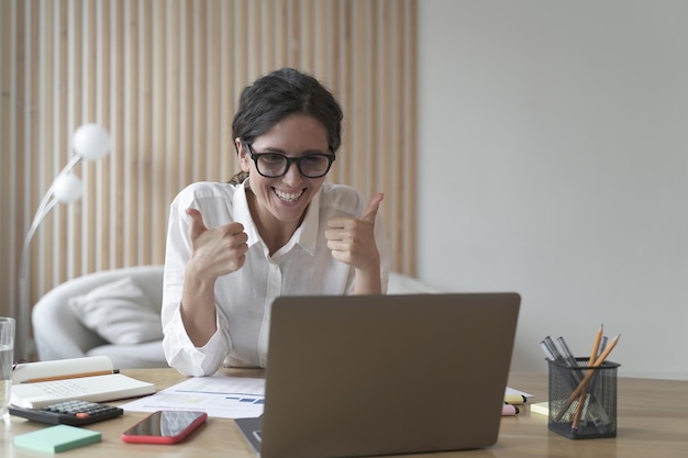 Mulher de negócios confiante olhando para a tela do computador com um sorriso largo enquanto mostra o polegar para cima com as duas mãos tutor feminino espanhol trabalhando no laptop no escritório em casa conceito de trabalho remoto