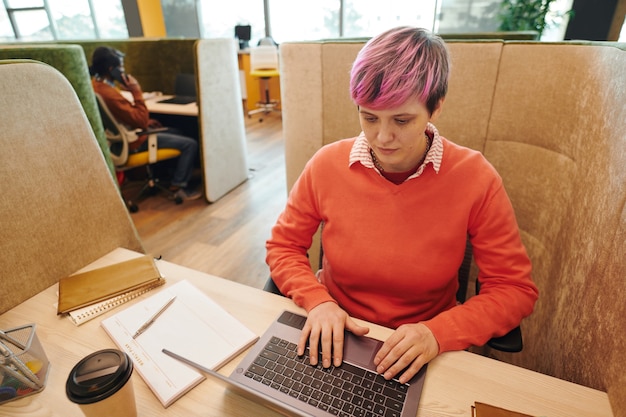Mulher de negócios contemporânea ou gerente de escritório em trajes casuais olhando para a tela do laptop enquanto está sentado na poltrona perto da mesa e rede