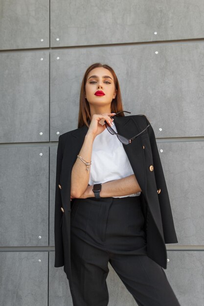 Foto mulher de negócios de beleza de moda com lábios vermelhos em terno elegante casual com óculos de sol está perto de uma parede cinzenta na rua