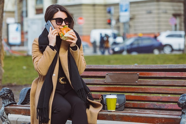 Mulher de negócios de beleza jovem comendo fast food e trabalhando no telefone enquanto está sentado no banco da cidade