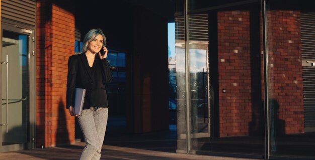 Mulher de negócios de cabelo azul andando na rua e discutindo no telefone segurando um laptop e sorrindo