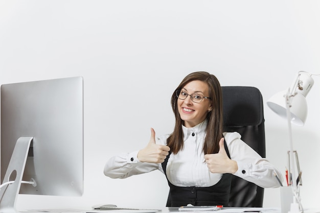 Mulher de negócios de cabelos castanhos muito sorridente de terno e óculos, sentada na mesa, trabalhando no computador com um monitor moderno com documentos em um escritório leve, mostrando o polegar para cima ambas as mãos