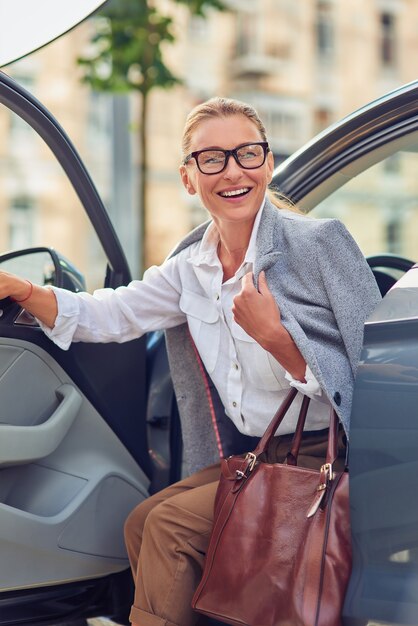 Mulher de negócios de meia-idade na moda bem-sucedida com bolsa saindo de seu carro moderno e