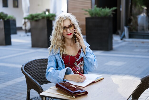 Mulher de negócios de óculos sentada em um café na rua, escrevendo em um caderno e falando ao telefone