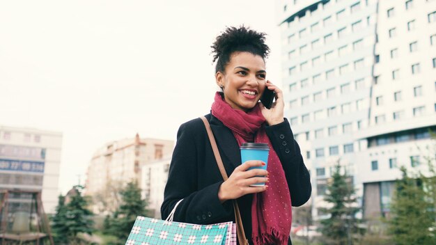 Mulher de negócios de raça mista atraente falando smartphone e bebendo café caminha na cidade