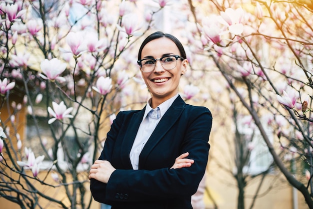 Mulher de negócios de sucesso feliz em óculos com braços cruzados olhando para a câmera no fundo da árvore de flores