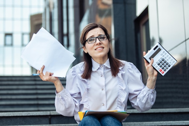 Mulher de negócios desanimada segurando uma calculadora e folhas de papel enquanto está sentada na escada Economist Lifestyle