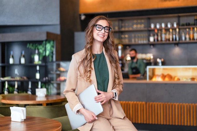 Mulher de negócios dona de restaurante com laptop nas mãos vestida elegante macacão em pé no restaurante com balcão de bar branco óculos feminino pessoa de negócios interna