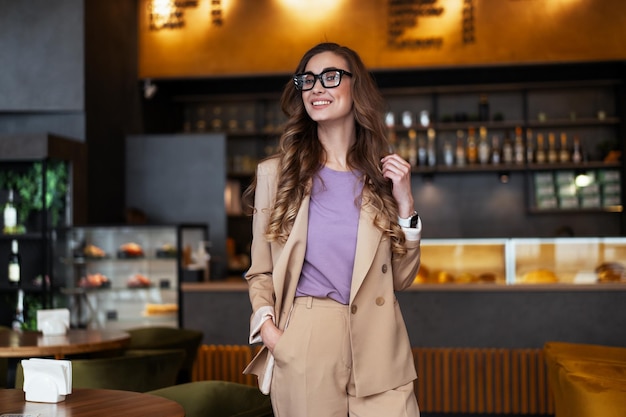 Mulher de negócios dono de restaurante vestido elegante terninho em pé no restaurante com balcão de bar óculos de mulher caucasiana pessoa de negócios interior mãos nos bolsos