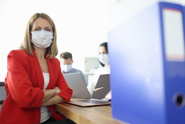 Mulher de negócios em um terno vermelho e máscara protetora no rosto se senta à mesa no escritório. modo de máscara de quarentena devido ao conceito de pandemia covid19.