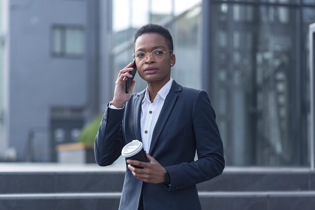 Mulher de negócios em um traje de negócios falando ao telefone, uma mulher afro-americana em um intervalo com um copo de bebida quente caminha no distrito comercial perto do escritório