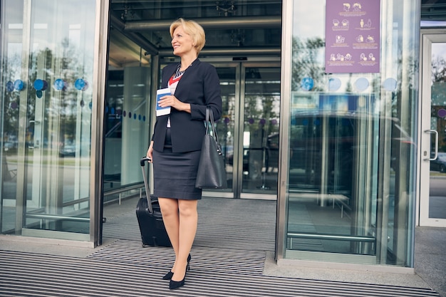 Mulher de negócios encantadora em um terno de saia elegante segurando uma mala de viagem e sorrindo enquanto sai do terminal do aeroporto