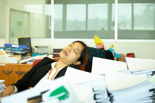 Mulher de negócios esgotada que dorme na mesa no escritório com a pilha do paperwok.